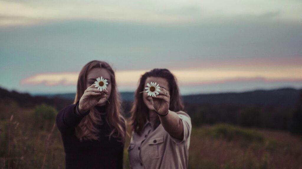 Two Women Outside At Dusk Smiling And Holding Daisies In Front Of Their Faces