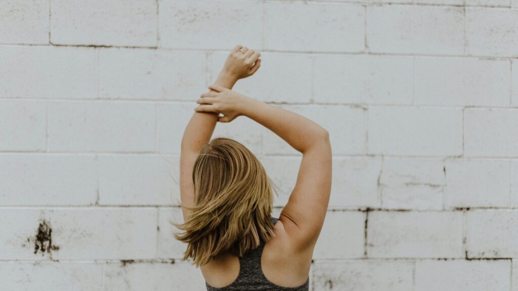 Girl In Workout Top With Arms Stretched Up In Front Of White Brick Wall