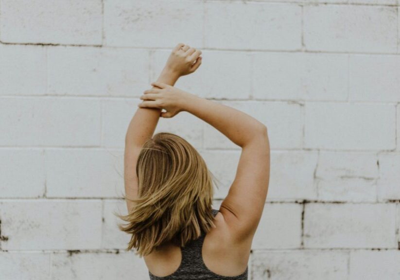 Girl In Workout Top With Arms Stretched Up In Front Of White Brick Wall