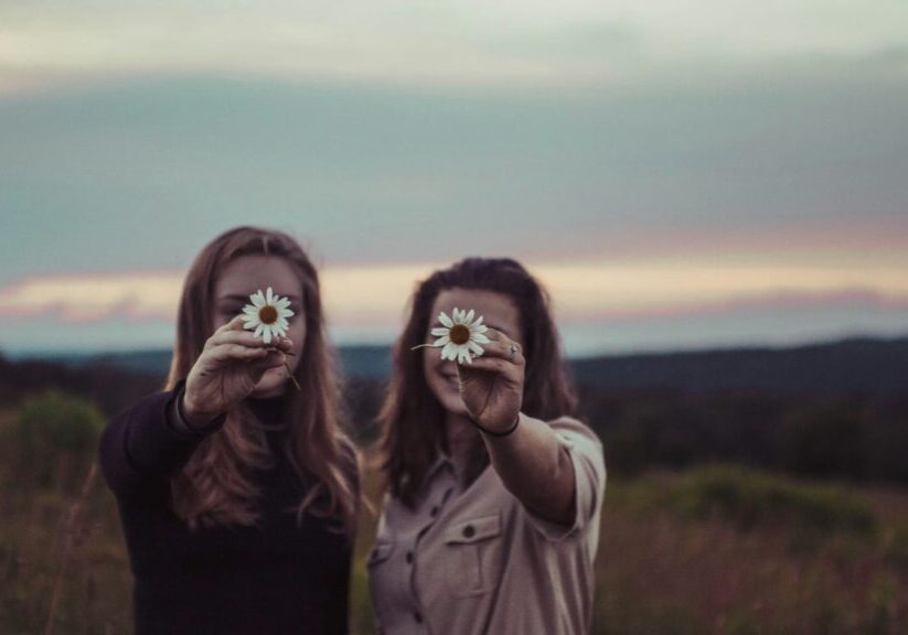 Two Women Outside At Dusk Smiling And Holding Daisies In Front Of Their Faces